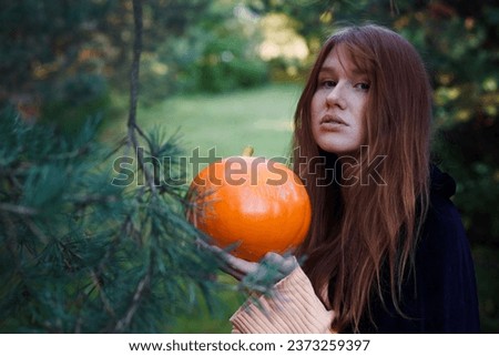 Witch young ginger woman in a black robe walks through the forest, the concept of Halloween, esotericism, mysticism, magic, sorcery, pumpkin
