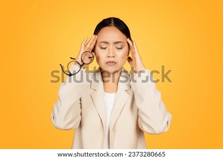 Asian woman, adorned in professional suit, endures moment of pain from migraine while at her work environment. Her expression reflects struggle of maintaining professionalism amidst discomfort