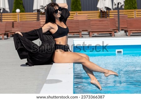 A girl in a black swimsuit and glasses sits by the pool.