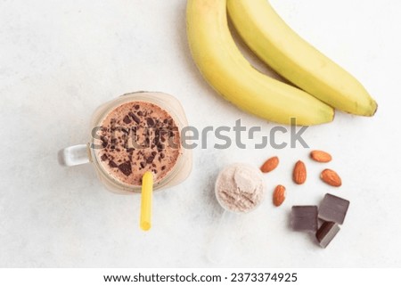 Glass jar of protein milkshake drink or smoothie and whey protein powder in measuring spoon, bananas, chocolate cubes, almond nuts on white background. bodybuilding sport nutrition, top view. Royalty-Free Stock Photo #2373374925