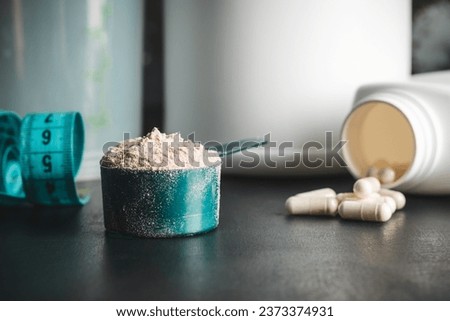 Chocolate whey protein powder in measuring spoon, white capsules of amino acids, vitamins and creatine, measuring tape, plastic shaker on dark background. bodybuilding food supplements. Royalty-Free Stock Photo #2373374931