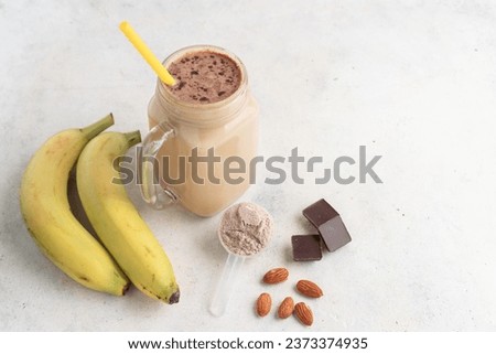 Glass jar of protein milkshake drink or smoothie and whey protein powder in measuring spoon, bananas, chocolate cubes, almond nuts on white background. sport nutrition, bodybuilding food supplements. Royalty-Free Stock Photo #2373374935