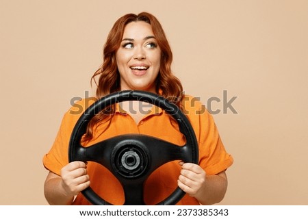 Young ginger chubby overweight woman wears orange shirt casual clothes hold steering wheel driving car look aside on area isolated on plain pastel beige background studio portrait. Lifestyle concept