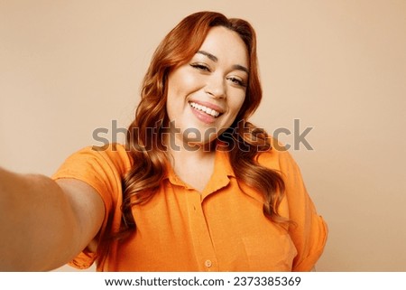 Close up happy young ginger chubby overweight woman wear orange shirt casual clothes doing selfie shot pov on mobile cell phone isolated on plain beige background studio portrait. Lifestyle concept