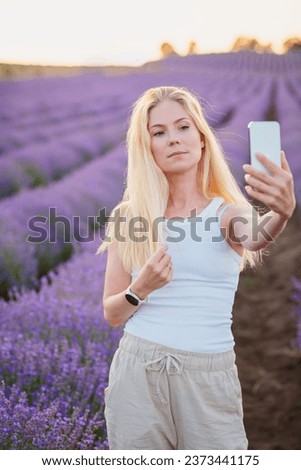 Lavender field. Pretty woman with blond hair walking between rows of lavender flowers with mobile phone, taking selfie, communicating in social networks, relaxing, resting, enjoying nature