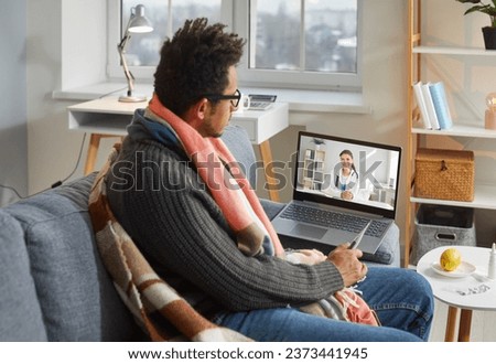 Sick man tells about his health problems to an online doctor. Young African American man with a fever sitting on the couch at home and having a remote telemedicine consultation on a modern laptop PC Royalty-Free Stock Photo #2373441945