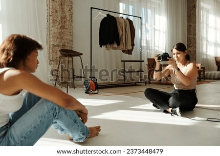 Young professional photographer sitting on the floor of spacious sunlit studio in front of female fashion model and taking photos of her