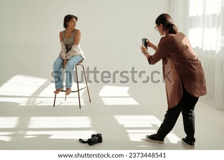Young fashion model sitting on chair and looking aside during photo session while female photographer taking picture on smartphone