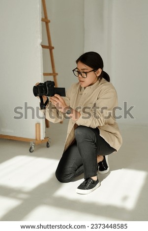Young woman with photocamera standing on knees on the floor and looking at screen during photo session before taking new shot of model