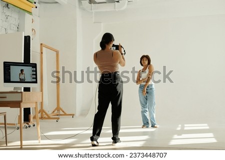 Long shot of young African American woman in casualwear posing for camera while standing in front of female photographer in studio