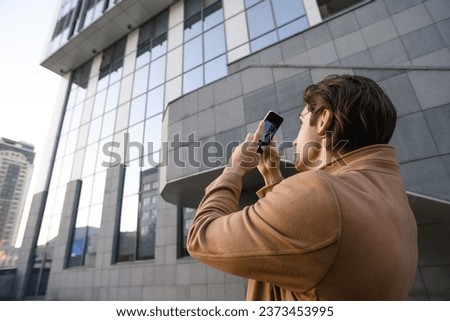 Brunette tourist in coat taking photo on smartphone on urban street at daylight	