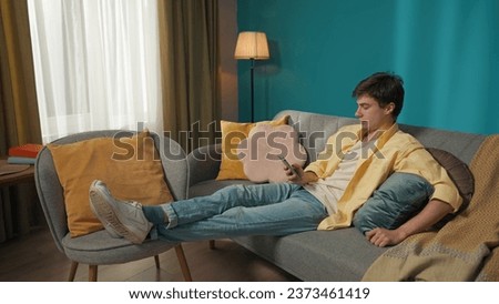Full-size shot capturing a young brunette man sitting on the couch, chatting with somebody, surfing online, scrolling his feed in social network.