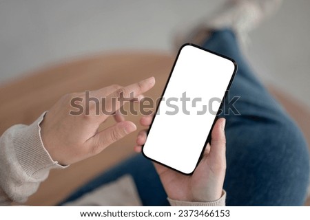 Top view image of a woman sitting indoors and using her smartphone to check messages or chat with her friends. A white-screen smartphone mockup in a woman's hand. Royalty-Free Stock Photo #2373466153