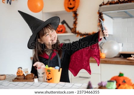 An adorable and joyful young Asian girl in a Halloween costume is taking selfies or her video with a smartphone in the kitchen, celebrating Halloween at home.