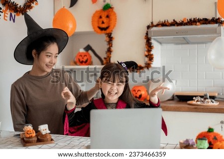 An overjoyed and happy young Asian girl in a Halloween costume enjoys watching Halloween cartoons on a laptop with her mom in the kitchen, celebrating Halloween season at home together.