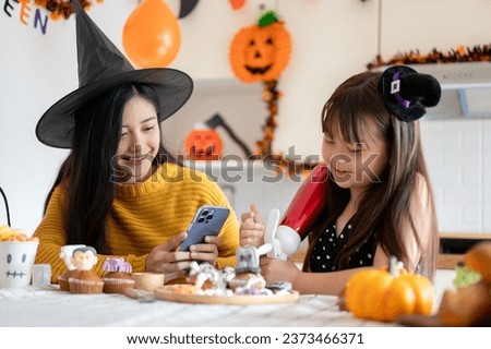 A kind and beautiful young Asian mom in a witch hat enjoys making Halloween cupcakes with her young daughter in the kitchen, celebrating Halloween at home together.