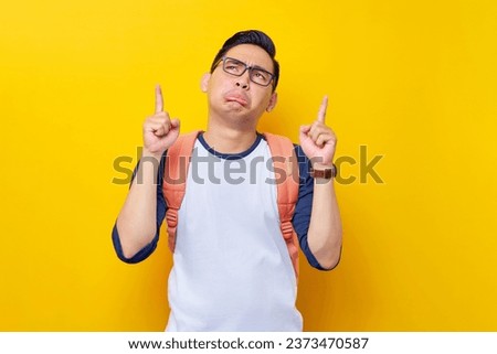 Frowning young Asian student man in casual clothes and glasses backpack pointing finger up at empty space isolated on yellow background. Education in High School University College concept