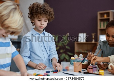 Portrait of cute curly haired boy coloring Easter pictures in preschool, copy space