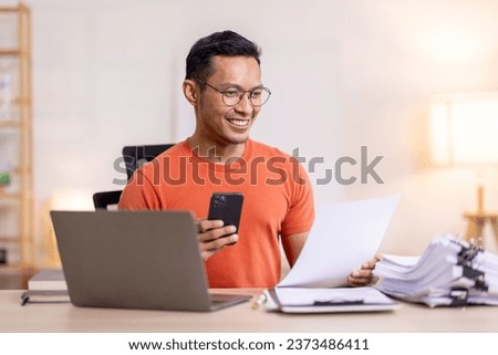 Picture of young asian man using a smartphone and smiling. working at home.accountant Documents data contract report for analysis TAX.