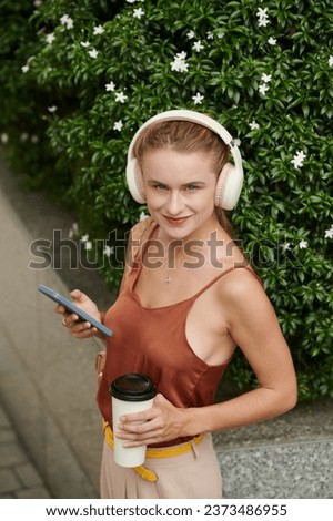 Portrait of smiling young woman with cup of coffee listening to music from playlist on smartphone