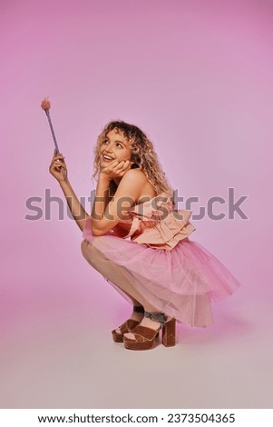 pretty woman in tooth fairy costume with magic wand in hands squatting and posing on pink backdrop Royalty-Free Stock Photo #2373504365