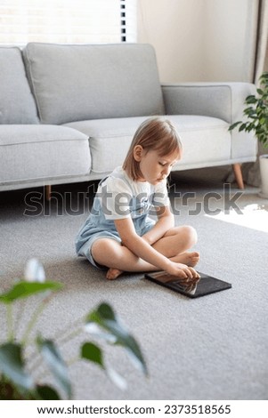 A cute preschool girl looks at the tablet screen while sitting on the floor in the room. Children's educational apps for phone and tablet. Children and digital technologies.