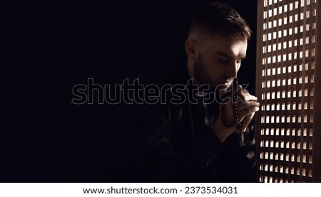 Young praying priest in confession booth Royalty-Free Stock Photo #2373534031