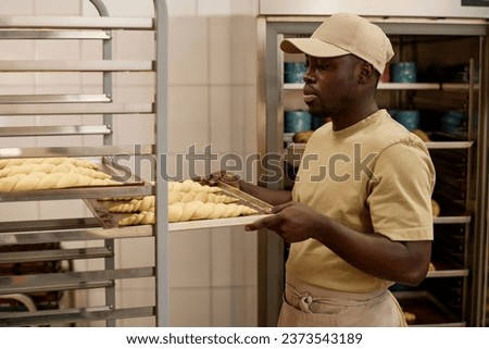 Side view portrait of Black young man preparing trays with fresh breads and buns while working in artisan bakery, copy space Royalty-Free Stock Photo #2373543189