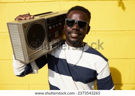Vibrant waist up portrait of funky Black man holding boombox outdoors and smiling at camera
