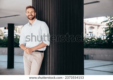 Handsome smiling stylish hipster lambersexual model. Modern man dressed in white shirt and trousers. Fashion male posing in the street urban background Royalty-Free Stock Photo #2373554153