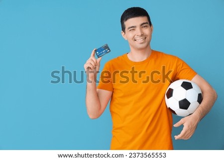 Happy young man with soccer ball and credit card on blue background. Sports bet concept