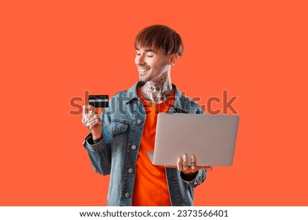 Tattooed young man with credit card and laptop on orange background