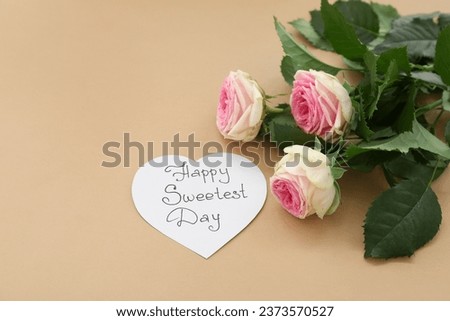 Paper heart with text HAPPY SWEETEST DAY and bouquet of rose flowers on brown background. National Sweetest Day Royalty-Free Stock Photo #2373570527