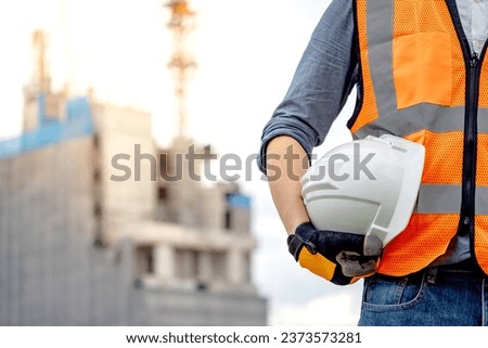 Safety workwear concept. Male hand holding white safety helmet or hard hat. Construction worker man with reflective orange vest and protective gloves standing at unfinished building with tower crane Royalty-Free Stock Photo #2373573281