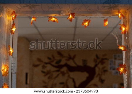 Autumn interior decor. Orange electric lights having maple tree leaves shape attached to room door frame. Fall tree painting on the wall seen from door. Halloween decoration ideas for kids home party.