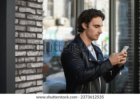 Stylish man in black leather jacket using mobile phone near building on urban street	