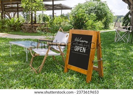 White wooden bench in a coffee shop with a message on a vintage chalkboard sign. "Welcome, we're open"  with beautiful green plant background.
