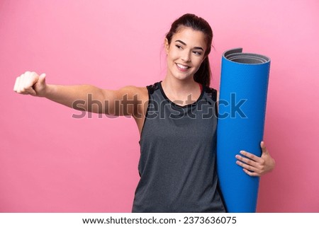 Young Brazilian sport woman going to yoga classes while holding a mat isolated on pink background giving a thumbs up gesture