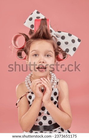 Pin-up style. A cute little girl in an elegant polka-dot dress and curlers on her head is going to a party. Pink studio background. Kid's Fashion - clothes and accessories.