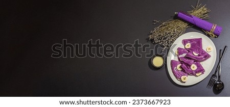 Thin lavender pancakes with banana slices and sweet topping. Healthy vegan non lactose and gluten free food, breakfast concept. Trendy hard light, dark shadow, black stone background, banner format