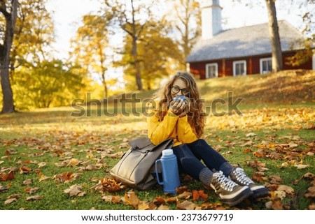 Beautiful tourist in a yellow raincoat with a backpack sits in a clearing among yellow fallen leaves, drinks tea from a thermos, enjoys the sunny weather in an autumn park. 