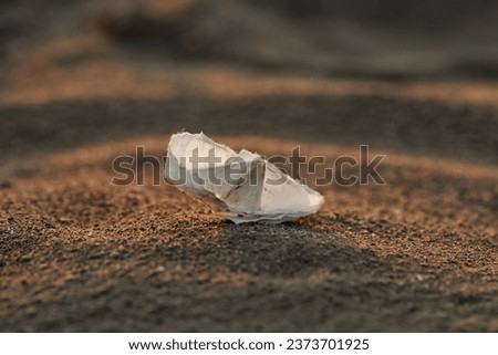 Closeup of a handmade white paper boat resting on a sandy beach, with a blurred oceanic landscape in the background
