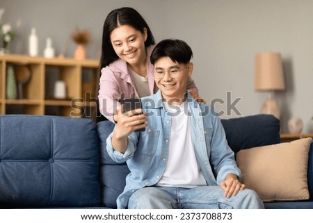 Smiling Korean couple engaging in online life with a smartphone, comfortably resting on sofa in modern living room at home. Relaxed, everyday technology use. Gadget and phone innovations