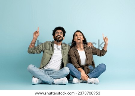 Loving cheerful young indian couple wearing casual outfits sitting on floor, looking up and pointing at copy space above for advertisement on blue studio background, full length. Great offer for two Royalty-Free Stock Photo #2373708953