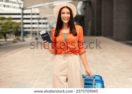 Cheerful woman traveler in hat with suitcase and tickets posing at airport outdoor, smiling looking at camera while waiting her flight. Joyful anticipation for vacation and travel adventures