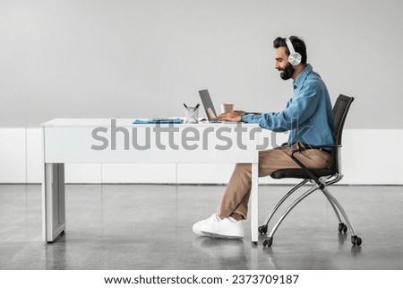 Side view of indian businessman in headphones working on laptop, making video call, sitting at desk, having web conference with customer in office, free space Royalty-Free Stock Photo #2373709187