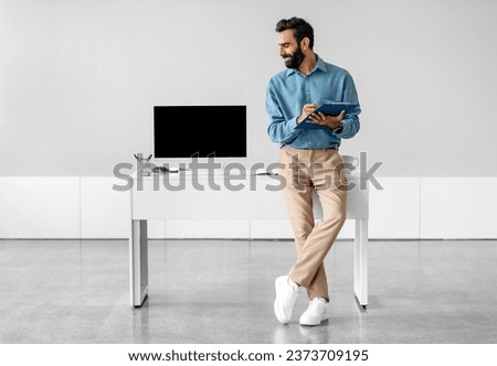 Confident indian businessman sitting leaning on table and looking at pc computer with blank black screen in office over light wall, free space. Technology concept Royalty-Free Stock Photo #2373709195
