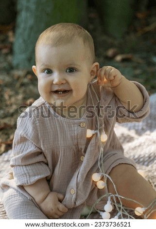 Portrait of an adorable little girl in a beige dress sitting near a large tree and looking at the camera. He holds lanterns and chews them with pleasure. Baby is one year old