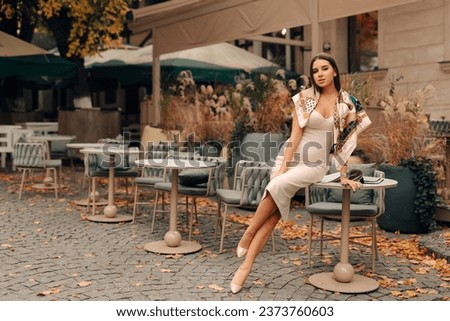 fashion outdoor photo of beautiful woman with dark hair in elegant dress posing in autumn street cafe