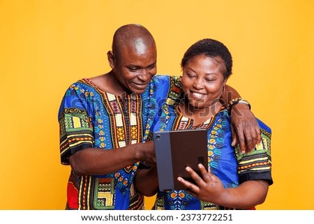 Smiling wife and husband with joyful expression hugging while using digital tablet together. Cheerful woman holding portable gadget while man tapping on touchscreen in studio